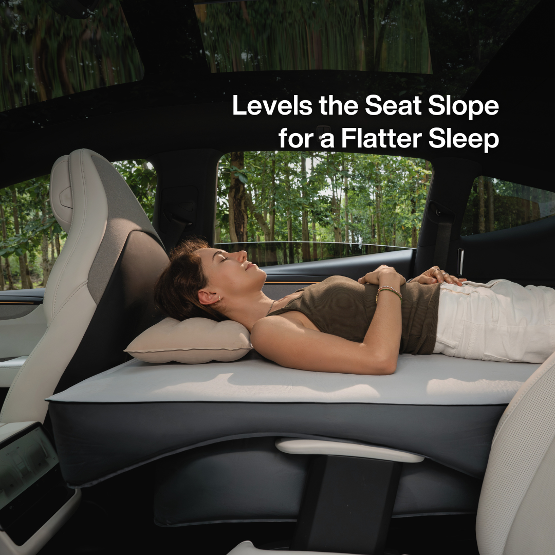 A woman lies on an elevated car bed mattress inside a vehicle, with text reading 'Levels the Seat Slope for a Flatter Sleep.' The scene shows the product in use within a real car interior and outdoor forest view, so this is Lifestyle rather than Product/Packaging.