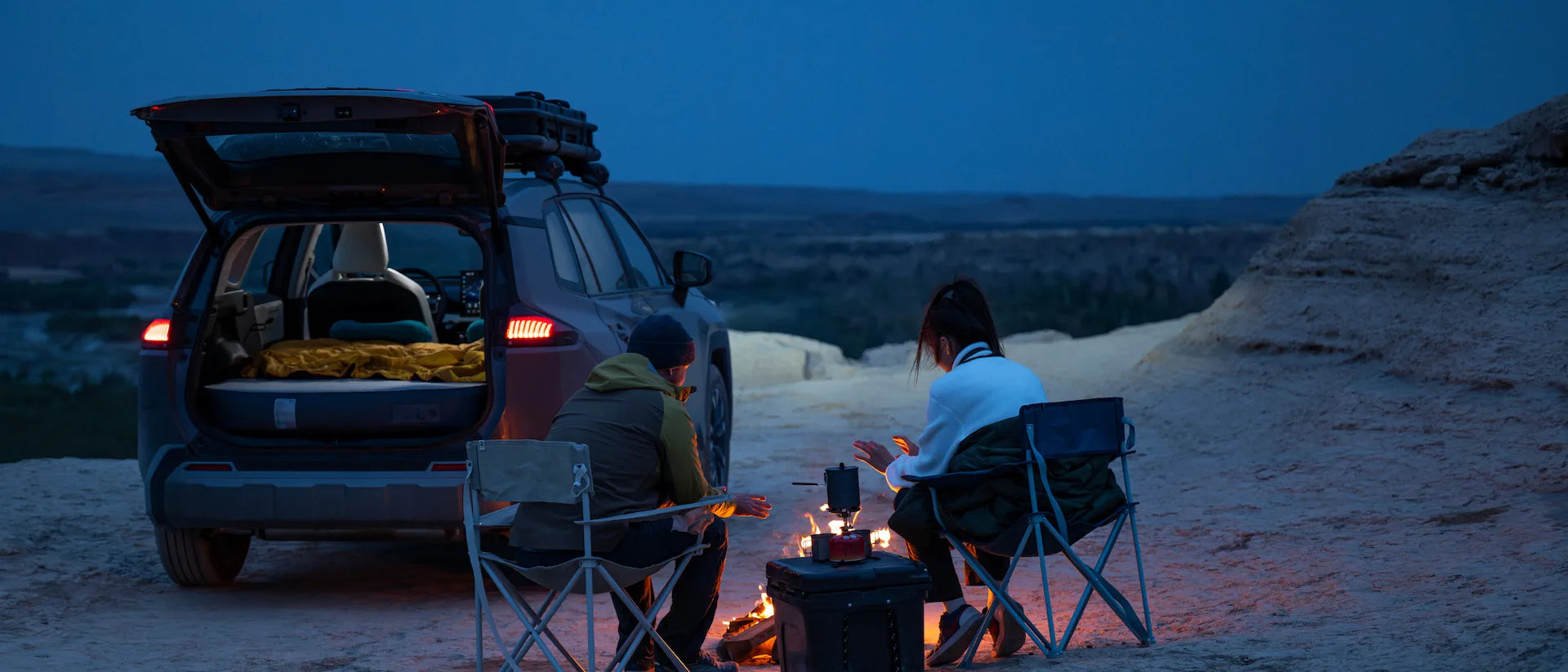 Two people car camping beside a vehicle with an open trunk at dusk